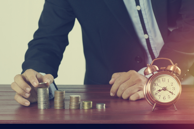 business man put money on pile of coins with alarm clock on working table concept in time to success